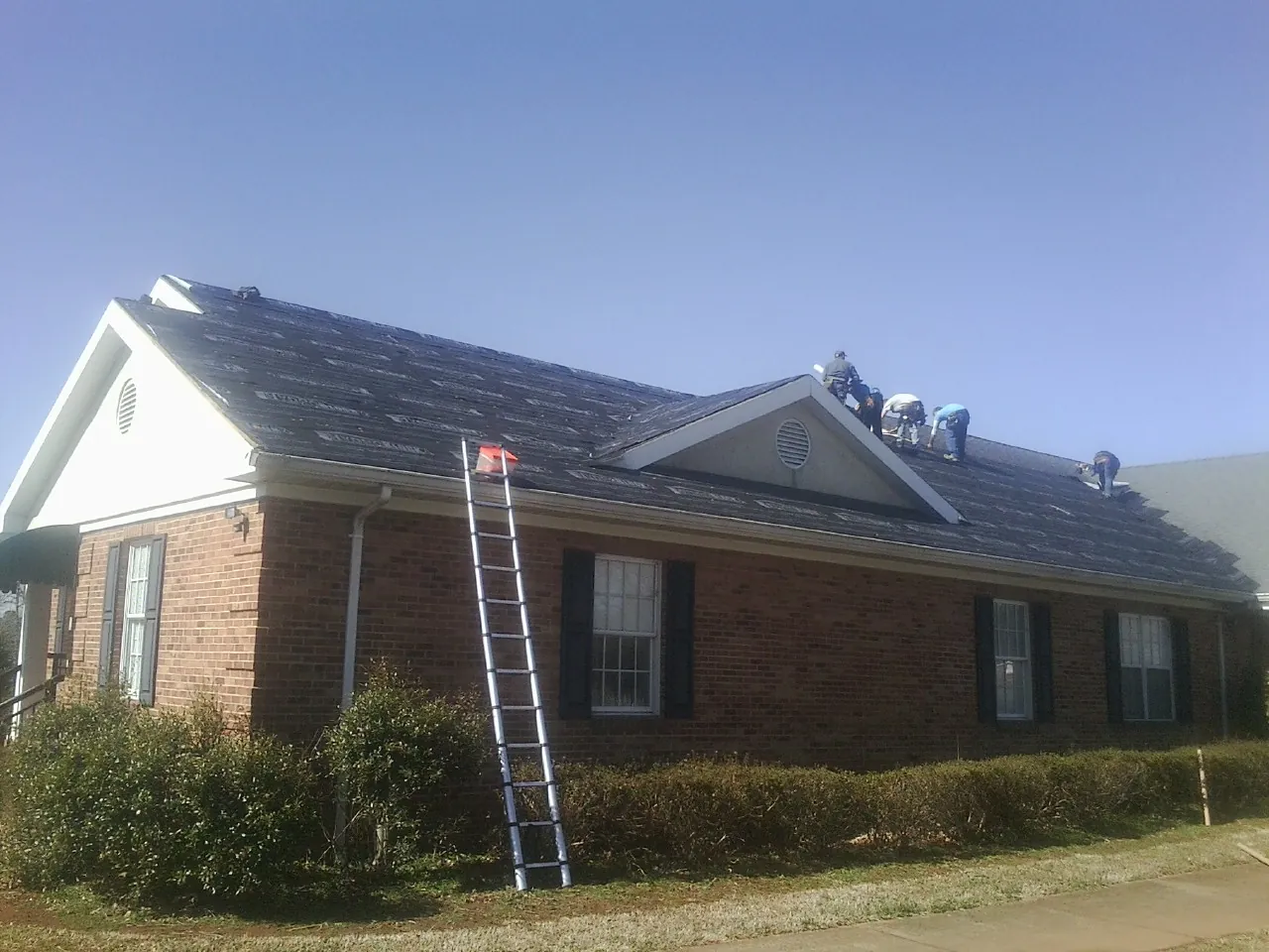 Crew tearing shingles off a brick ranch roof with a ladder set — Metal Roof Repair work by our crew in Ravena