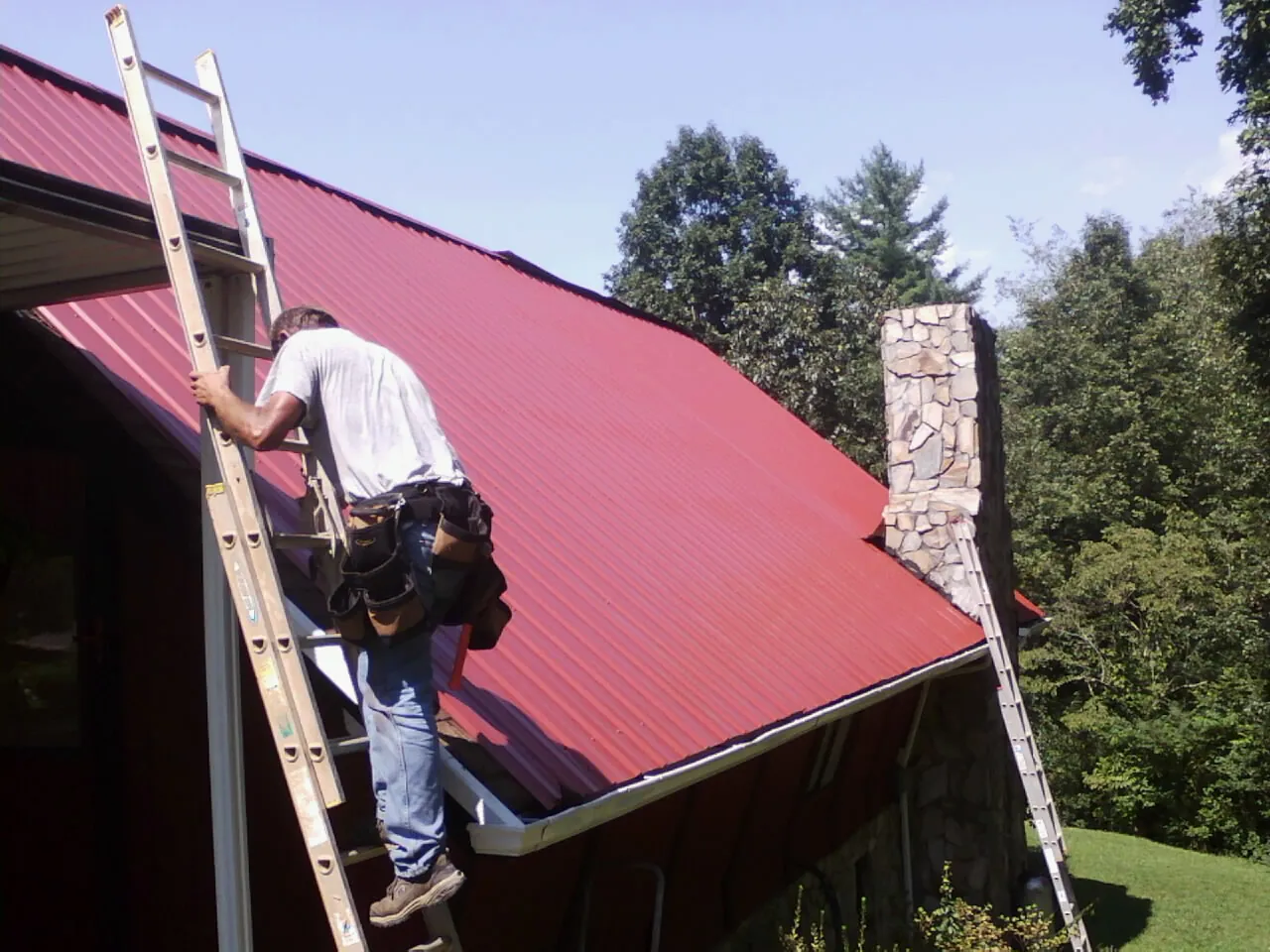 Roofer on a ladder beside a red metal roof and stone chimney — Soffit & Fascia Repair by Roofing Brothers in Ravena